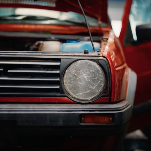 Gros plan sur l'avant d'une Volkswagen Golf de première génération rouge et noire avec capot ouvert, phare rayé et logo VW visible sur la calandre chromée. Le véhicule vintage illustre la campagne Volkswagen et DDB Paris sur le recyclage automobile joyeux, transformant la fin de vie des voitures en ode à l'économie circulaire avec une approche rock'n'roll et décalée.